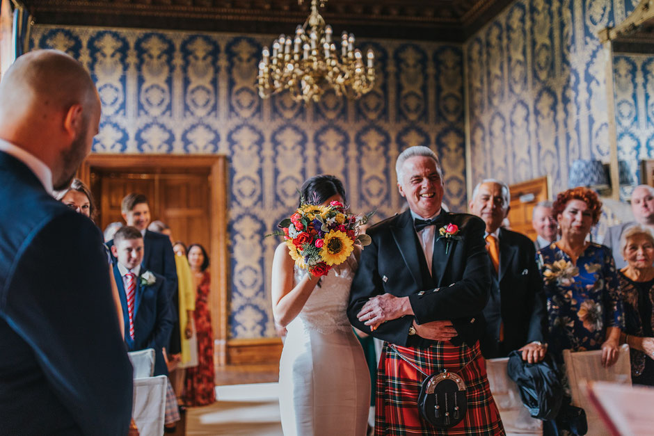 A shy bride covering her face with a colourful bouquet as she walks down the aisle