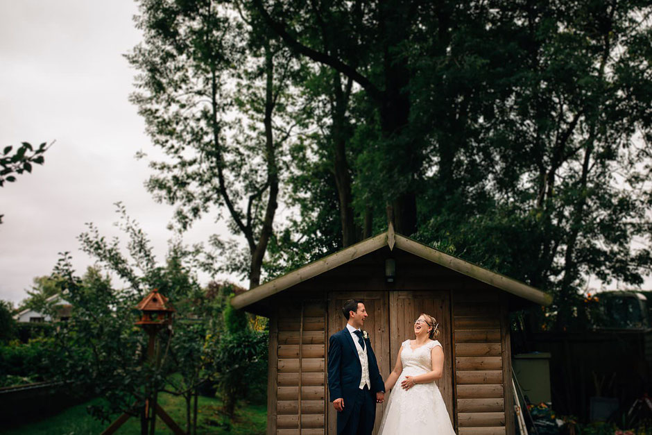 bride and groom laughing whilst stood infant of a wooden shed