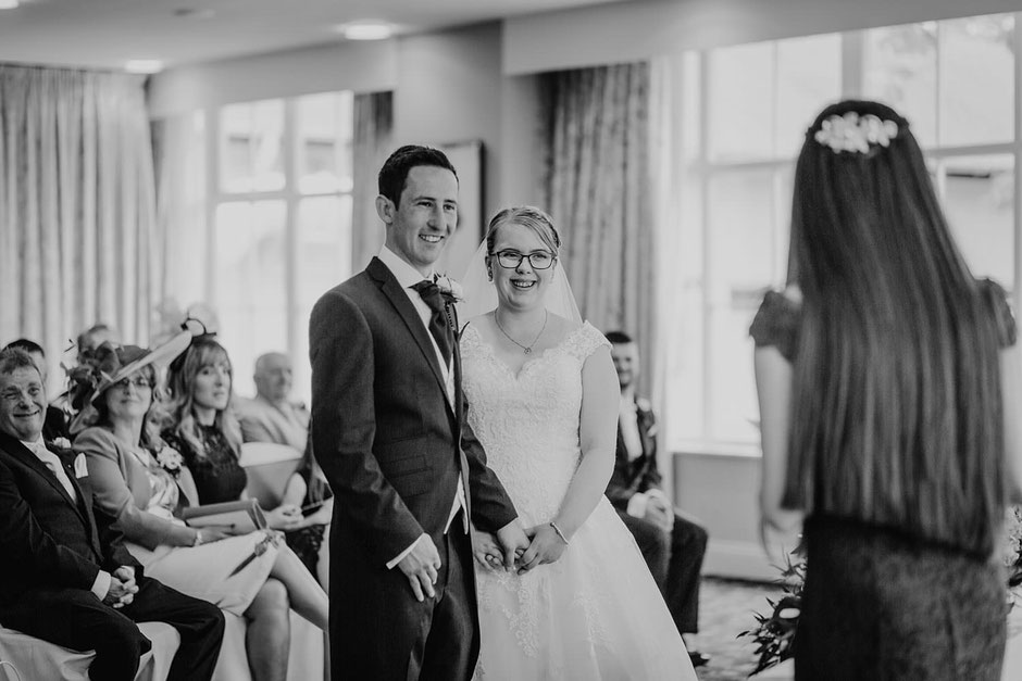 black and white photograph of a bride and groom listening to a reading by a bridesmaid during the wedding ceremony