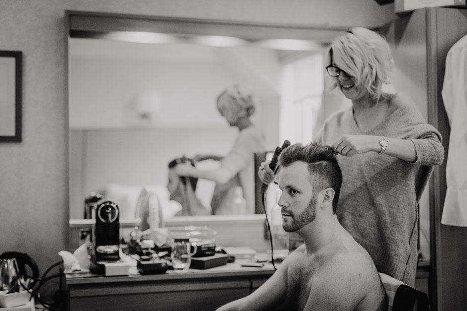 Black and white photo of a groom having his hair styled on the morning of his wedding day