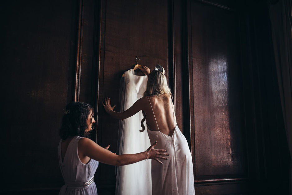 two ladies, one stood on a chair reaching for a wedding dress that is hung on a wall