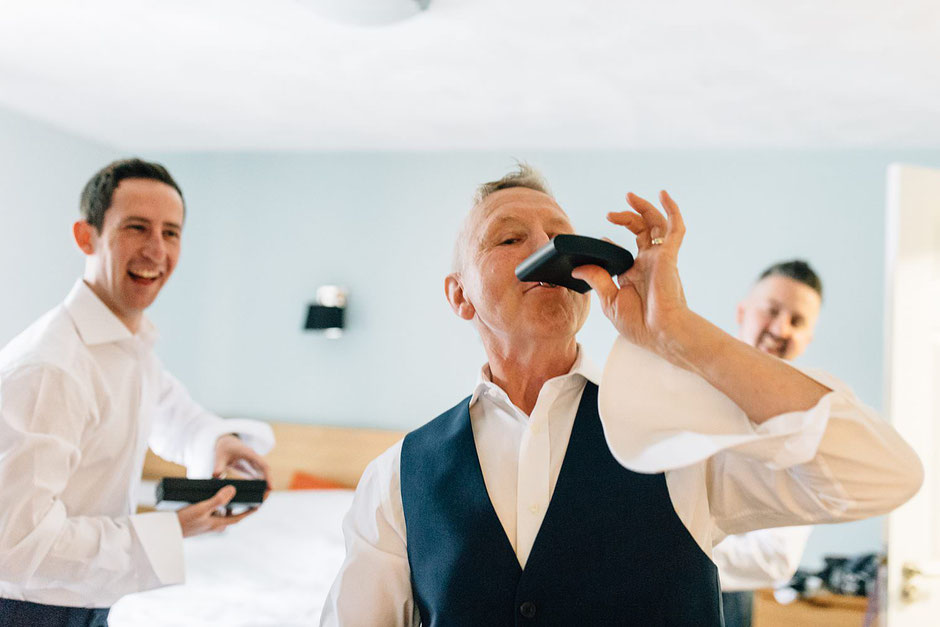 a man drinking out of a hip flask whilst two men laugh behind him