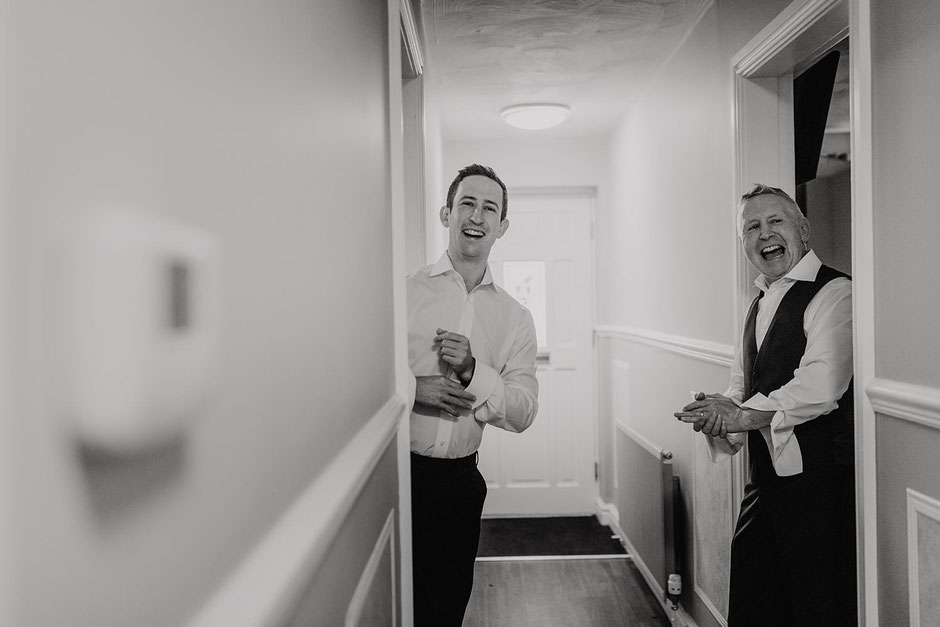A black and white photograph of a groom and his dad in a corridor laughing