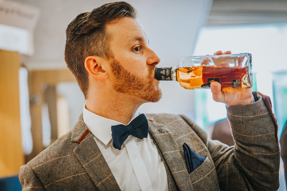 Groom dressed in a tweed suit with bowtie drinking straight from a bottle of whiskey