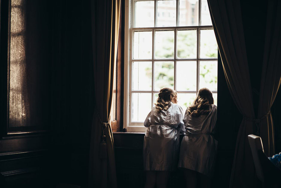 two bridesmaids dressed in silk dressing gowns looking out of a tall window