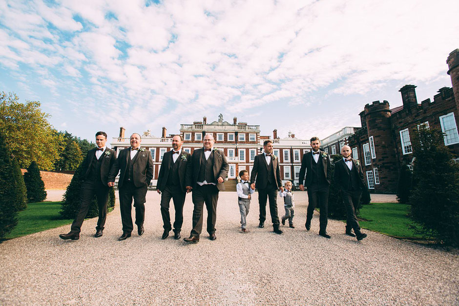 Grooms party in tweed suits walking down the driveway in front of Knowsley hall