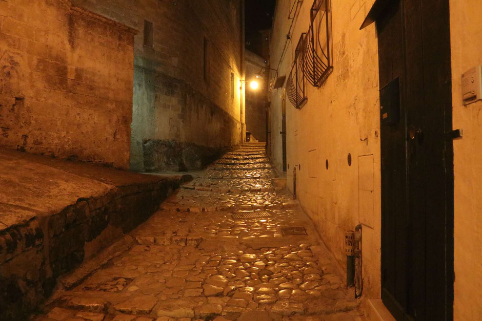 Menschenleere Gasse mit blank gelaufenen, unregelmäßigen Steinen im warmem Licht der Straßenlaternen am Abend in Matera ©My own Travel