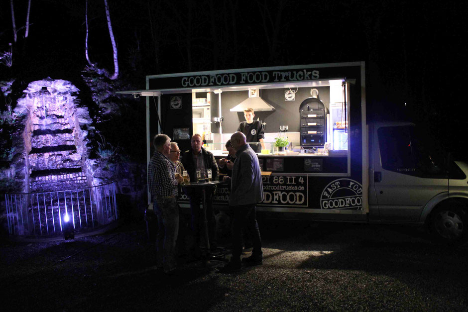 Gruppe am Stehtisch vor dem GOODFOOD FOOD TRUCK, Nachtaufnahme mit beleuchtetem Brunnen im Hintergrund des Food Trucks