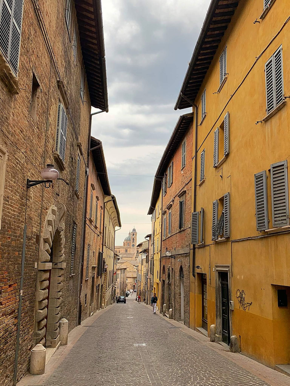 Street in Urbino with colored facades and in the distance the Palazzo Ducale, well worth a visit from your holiday accommodation Vecchi Maestri in San Pietro, Ancona. Adults only holiday apartments in Le Marche with pool and gym. Book now.