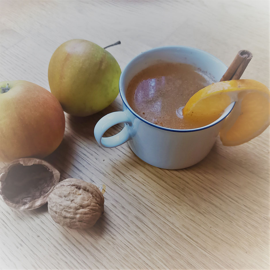 Tasse mit Getränk, Zimtstange, Zitronenscheibe und Äpfel zur Dekoration auf einem Tisch.