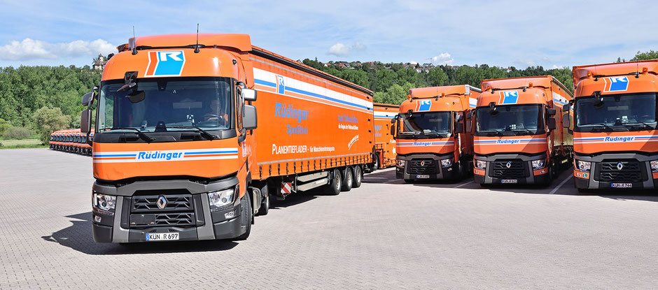 Eine lange Reihe an Lkw mit Anhängern ist ordentlich aufgereiht und wird zum Hintergrund hin immer kleiner. Man sieht von vorne auf sie. Aus dieser Auftstellung fährt ein Lkw hinaus und auf den Betrachter zu. Im Hintergund ist Wald.