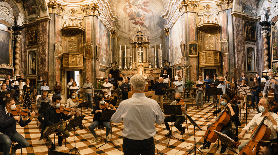 Dirigent im weißen Hemd steht vor seinem Orchester. Vorne Streichinstrumente, im Hintergrund ein Chor. Das Ganze spielt in einer Kapelle.