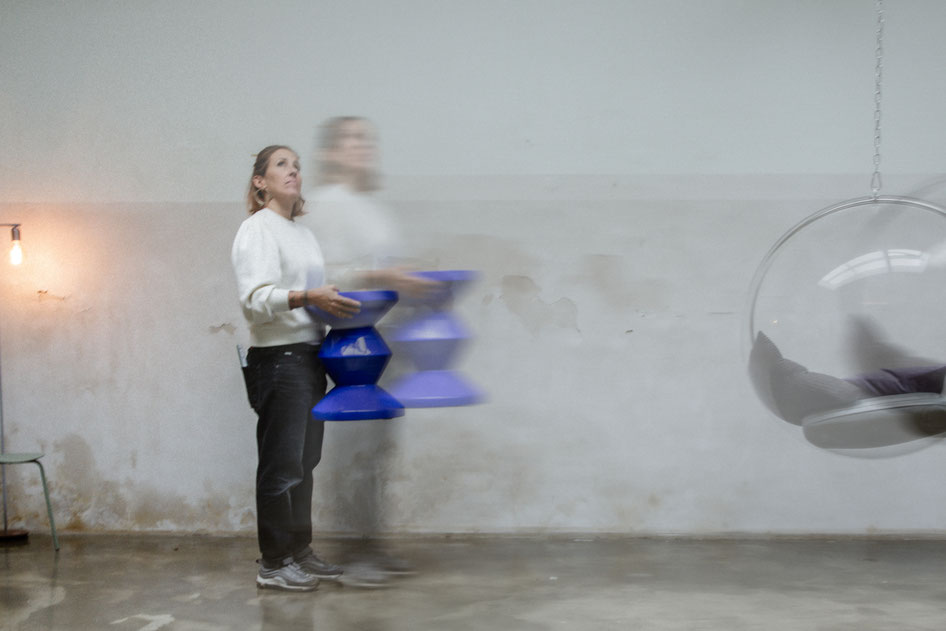 eine Frau mit sportlichen Schuhen steht in einem modernen Fotostudio und hält einen blauen Hocker Richtung Himmel Es geht dabei um Umzüge