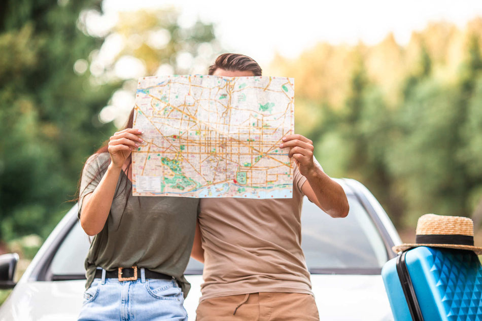 Paar mit Landkarte vor dem Gesicht steht vor einem Auto mit einem blauen Koffer