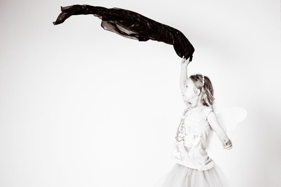 Schwarz-weiß-Fotografie eines kleinen Mädchens mit Engelsflügeln, welches mit einem Tuch winkt