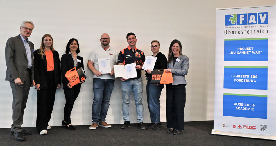 Zertifikatsverleihung mit mehreren Personen auf einer Bühne. Drei Teilnehmer präsentieren stolz ihre Auszeichnungen. Links und rechts stehen Vertreterinnen und Vertreter des FAV OÖ mit orangefarbenen Geschenktaschen.