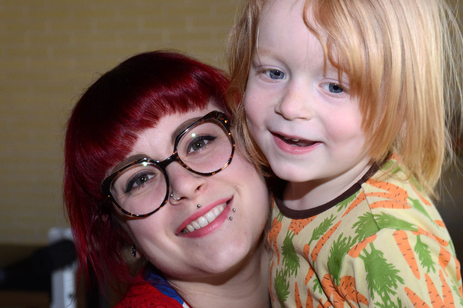Porträt Anke, rothaarig mit Brille und Piercings mit Noah im T-Shirt mit Karottenmotiv auf dem Arm