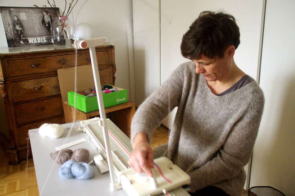 Eva im grauen Strickpulli sitzend rechts im Halbprofil arbeitet an Strickmaschine, die vor ihr steht