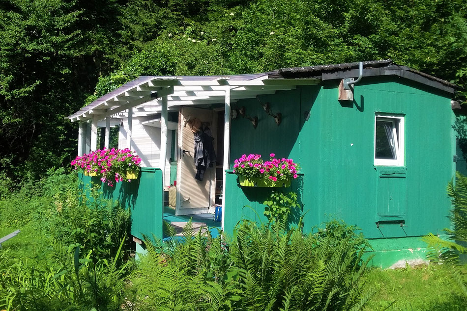 eine grün gestrichene Holzhütte. Davor eine Veranda mit weißen Pfosten und pinken Petunien n Blumenkästen. Um die Hütte Garten