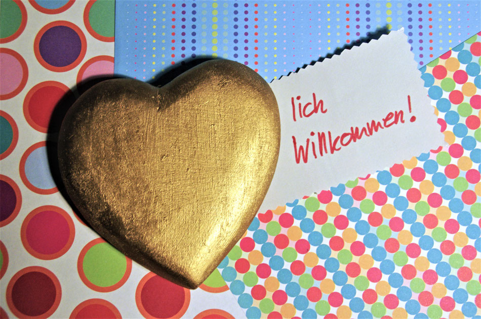 goldenes Herz auf bunt gemustertetem Untergrund. Neben dem Herz Schrift "lich Willkommen!"