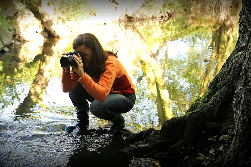 Sybille hockt mit Gummistiefeln in einem Bach und fotografiert