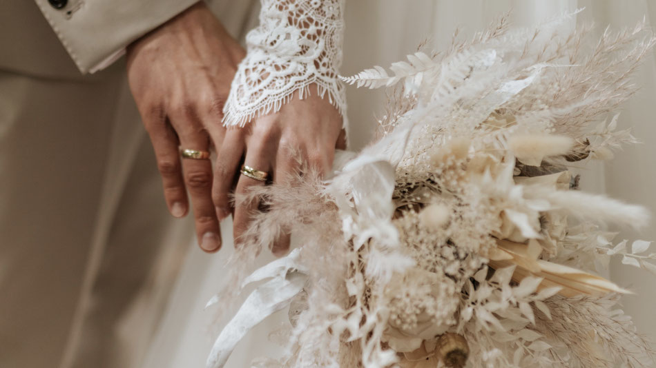 Ein Strauss Trockenblumen, dahinter sind zwei Hände zu sehen mit Goldringen die glänzen