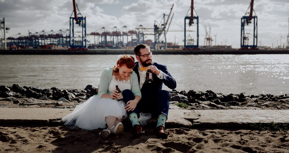 Ein Brautpaar sitzt lachend am Elbstrand. Im Hintergrund sind die Kräne des Hamburger Hafens zu sehen – freie Trauung mit Constanze Marienfeld.