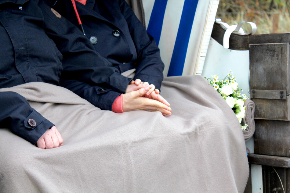 Ein Paar sitzt händestreichelnd im Strandkorb mit einer Decke. Seitlich liegt ein Strauß Blumen