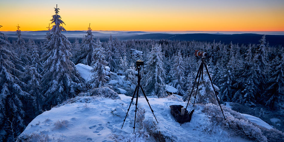 Morgendämmerung im winterlichen Fichtelgebirge. Auf verschneiten Felsen stehen zwei Foto-Stative, ausgerichtet auf den farbigen Sonnenaufgang. Der Rauhe Kulm ist zu sehen.