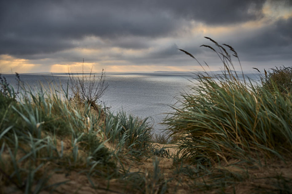 Blick durch Dünen mit Gras auf ruhiges Meer unter bewölktem Himmel bei Sonnenuntergang