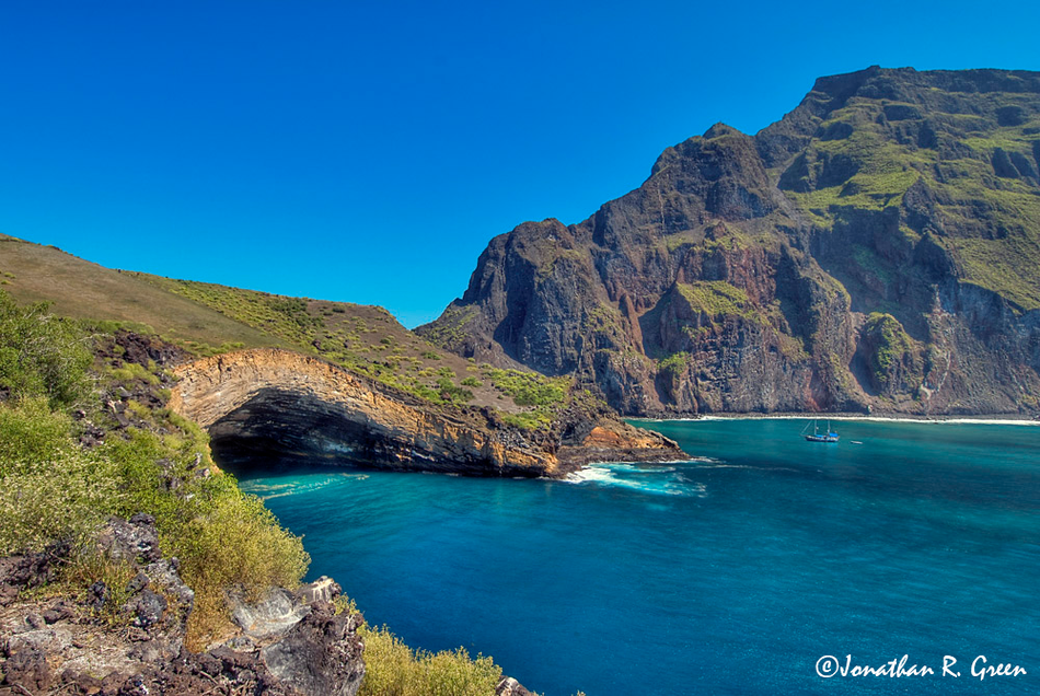 A coastal landscape view of the Pirate Cave at the Galapagos Islands, showing a large, naturally formed cave archway at the base of a steep, rugged cliff. The cliff itself displays a variety of green vegetation at the top.