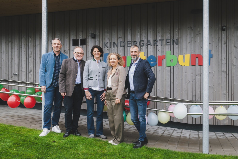 Fünf Personen stehen lächelnd vor dem Kindergarten „Kunterbunt“ in Zell am See-Schüttdorf. Hinter ihnen sind bunte Buchstaben und Luftballons zu sehen – ein fröhlicher Moment zur Eröffnung des neuen, familienfreundlichen Betriebskindergartens.