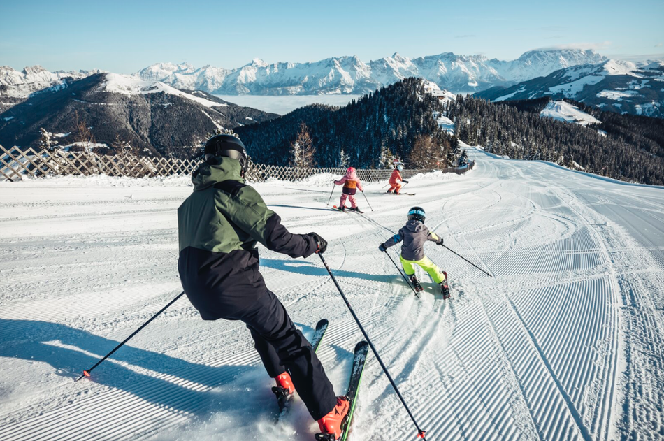 Kinder fahren bei besten Bedingungen eine sonnige, gut präparierte Skipiste in den Bergen hinunter – beste Bedingungen für große und kleine Wintersportfans.
