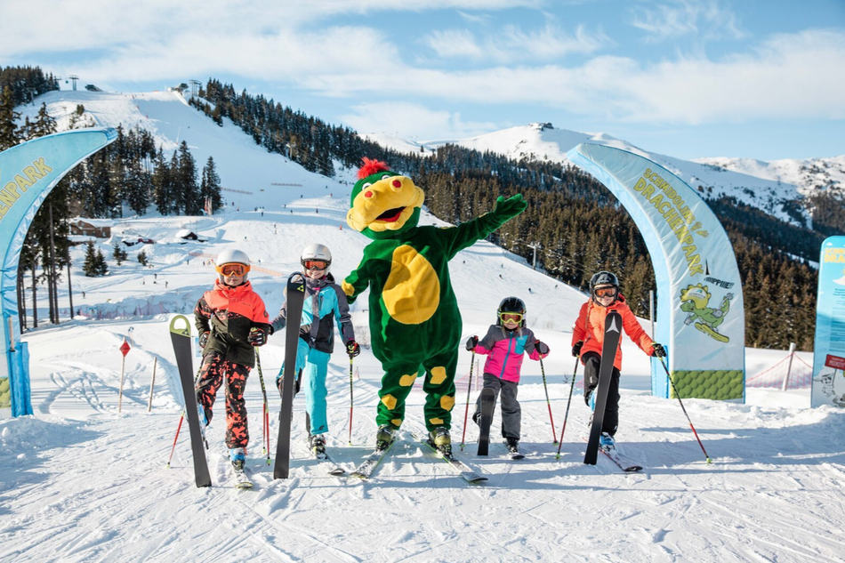 Kinder posieren fröhlich mit dem Maskottchen Schmidolin im Drachenpark auf der Schmittenhöhe – ein buntes Wintersporterlebnis bei Sonnenschein und Pulverschnee.