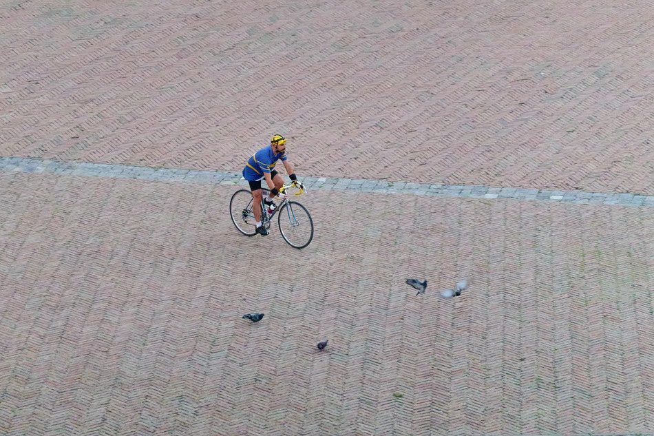 The image shows a photograph of a racing cyclist with vintage gear crossing an empty square with some pigeons.