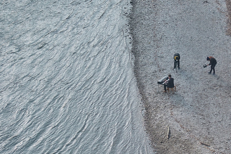 The image shows the aerial photograph of a piano player on the shore of the Aare river in Bern. There are two men filming the piano player.