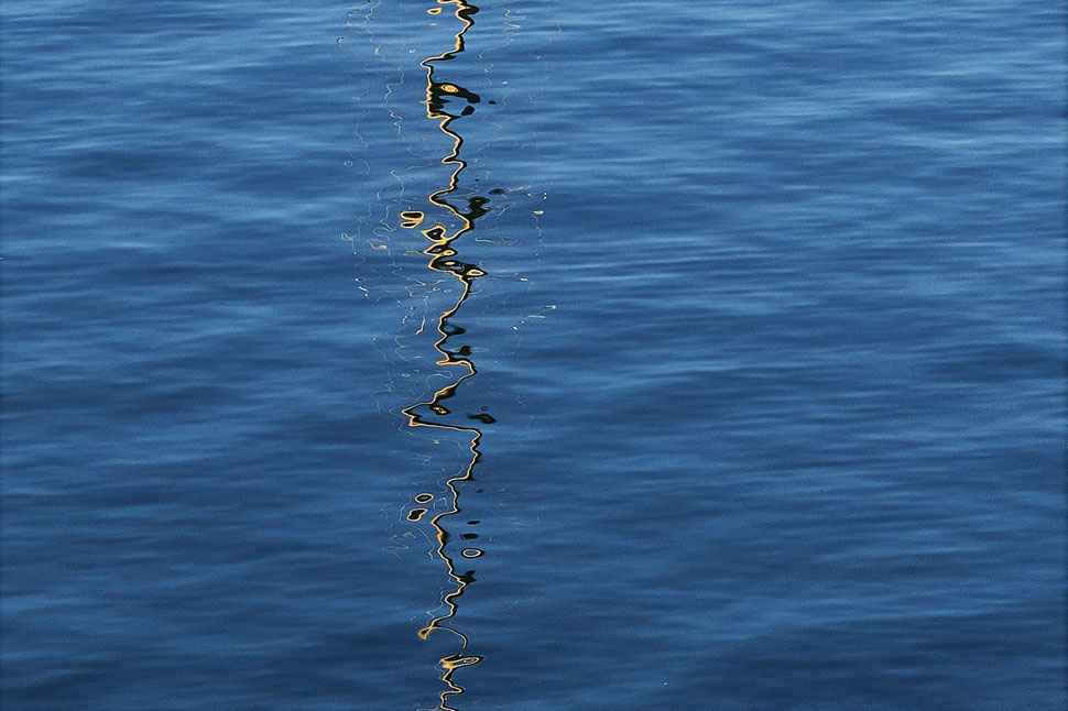 The image shows the photograph of lake water with the distorted reflection of a mast of a sailboat in the morning light.