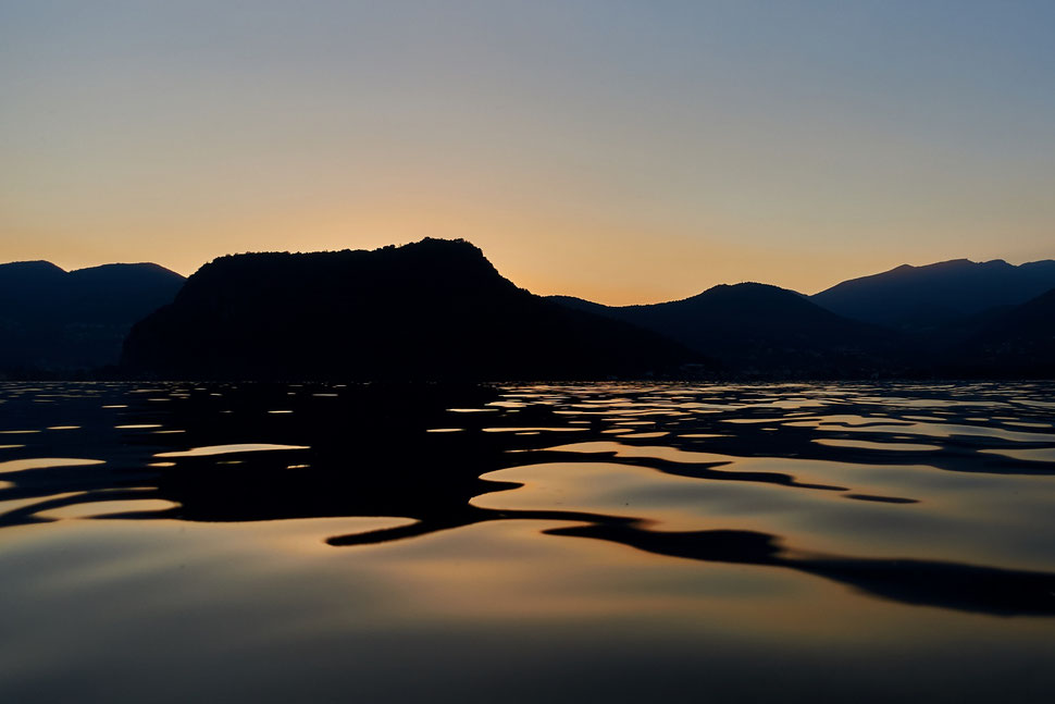 The image shows the photograph of light and shadow patterns in the water of Lake Lugano. It is after sunset and you can see the dark shapes of mountains in the background.