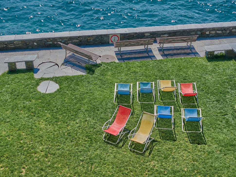 The image shows a photograph of 8 deck chairs with different colors on a lawn and three benches near the shore of a lake.