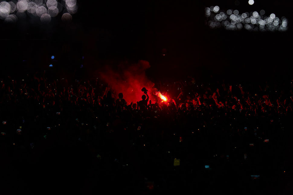 The image shows the night photograph of a large crowd at a music festival lit by a pyro flare. 