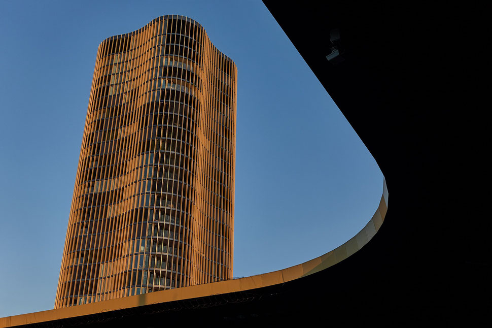 The image shows the photograph of a snippet of a high-rise building and part of the roof of Lucerne's football stadium. The high-rise building is illuminated by late afternoon sunlight.