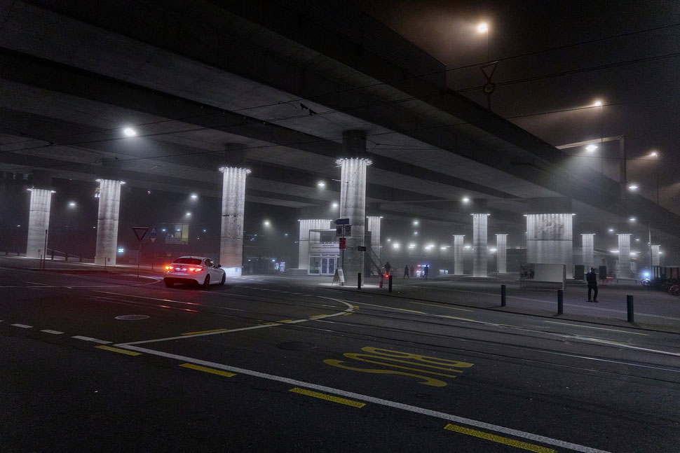 The picture shows a night photograph of a bridge with big pillars. A car turns around and drives under the bridge.