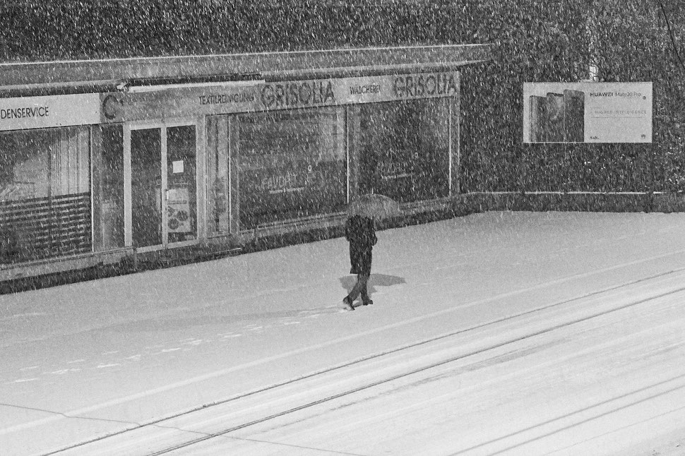 The image shows the photograph of a man with an umbrella walking on a snow covered street. There is a storefront next to him.