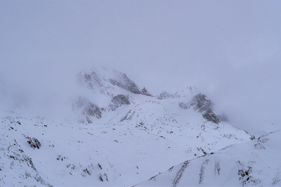 The image shows the photograph of faint shapes of mountains with snow agains the backdrop of a white sky.