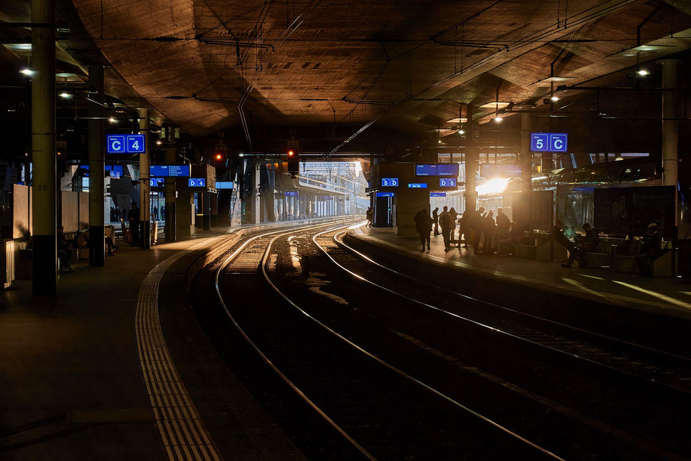 The image shows the photograph of people waiting at the train station in Bern while the sun is setting shining its beautiful light on the platforms. 