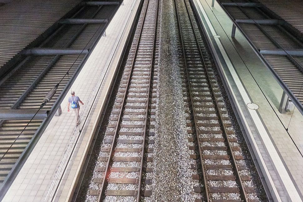 The image shows the photograph of an aerial view of a man wallking along train tracks at a train station late at night. The platform is empty.