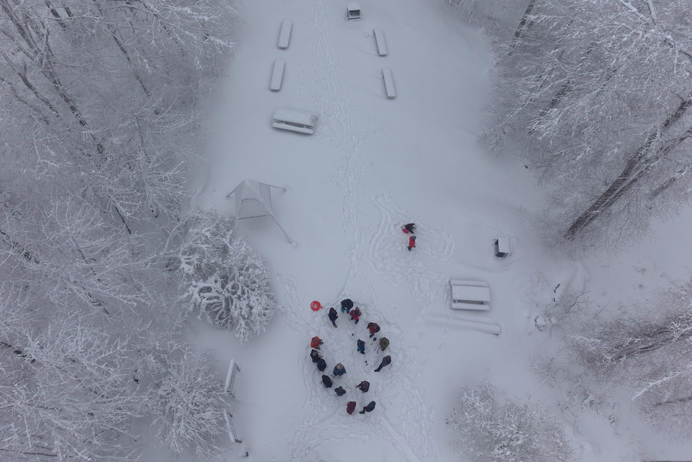 The image shows the aerial photograph of a group of people standing on the snow covered ground of a clearing.