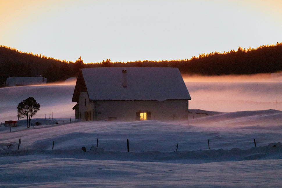 The image shows the photograph of a farm house in a Winter landscape of the Jura region (Switzerland). It is a very cold evening - the last sunlight illuminates the haze surrounding the house.