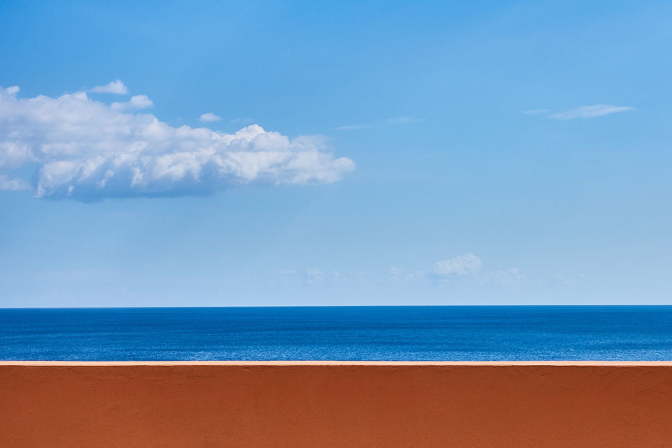 The image shows the photograph of a brown wall against the background of the dark blu sea and light blue sky with some clouds. 