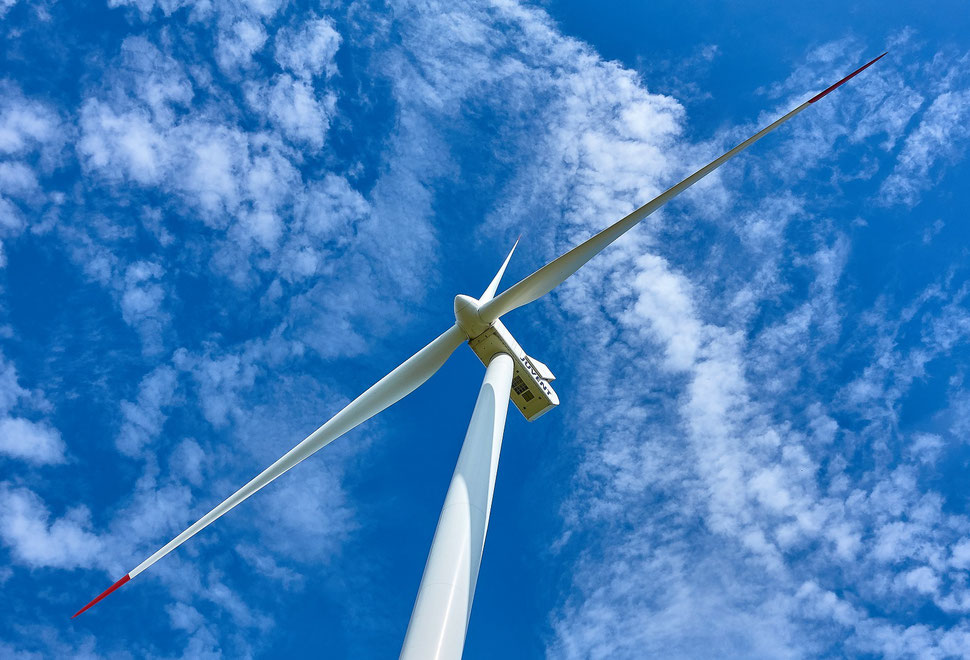 The image shows the photograph of a wind turbine from below against the backdrop of a blue sky with white clouds. 